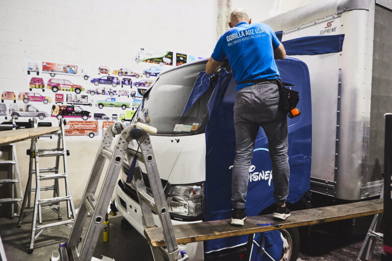Person applying Disney branding to a mobile billboard truck in a workshop, surrounded by marketing materials and ladders, showcasing preparation for the Disney+ activation campaign.