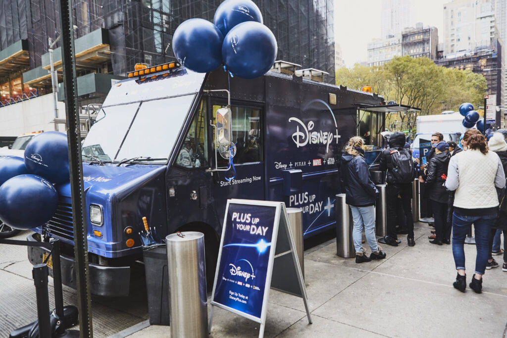 Disney+ promotional food truck with blue balloons and a sign stating "PLUS UP YOUR DAY" in Manhattan, NY, showcasing a guerrilla marketing campaign.