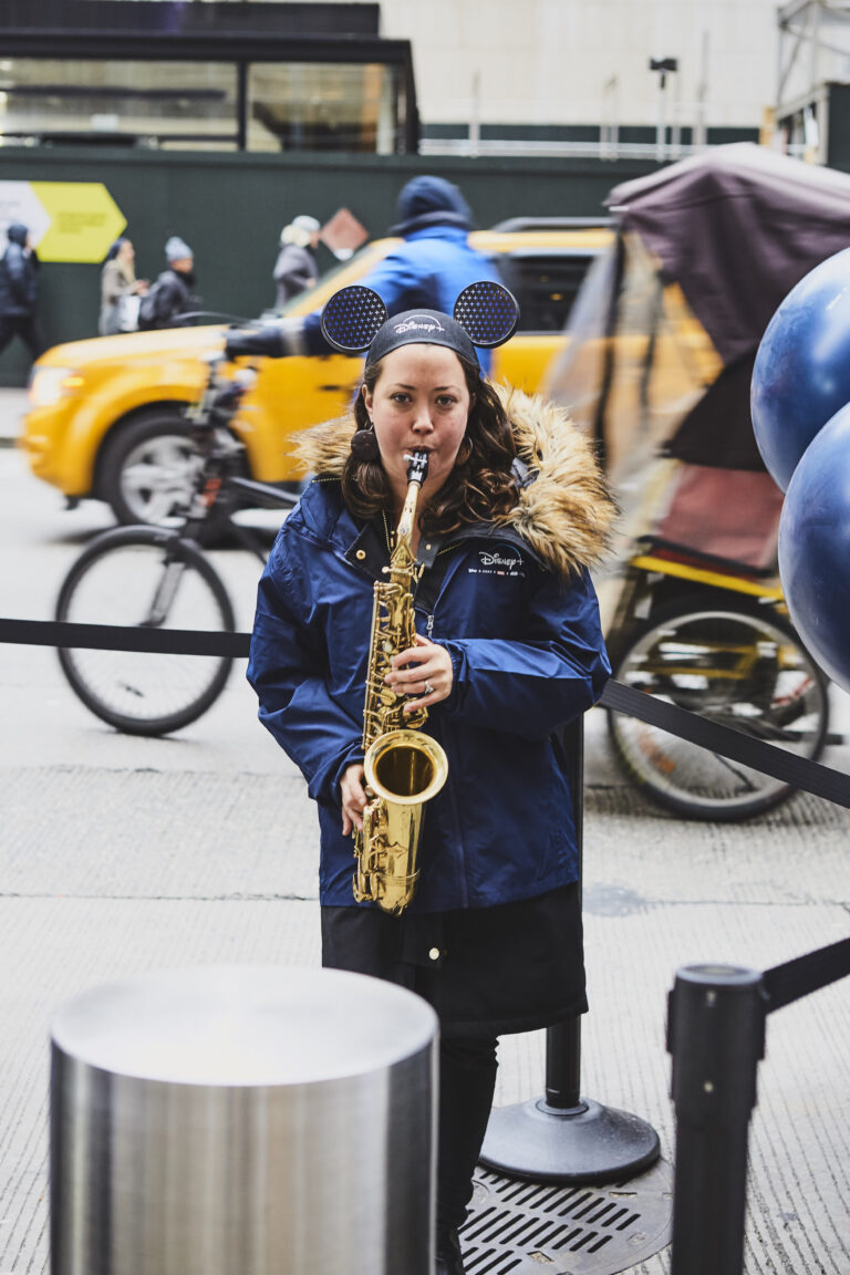 Saxophonist wearing Disney+ branded jacket and Mickey Mouse ears performing in Manhattan during Disney+ activation event, with yellow taxi and pedestrians in background.