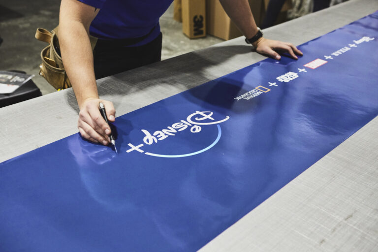 Person writing on a blue banner featuring the Disney+ logo, preparing for a marketing activation event related to the Disney+ launch in New York City.