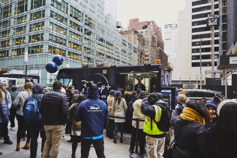 Crowd gathered around Disney+ mobile billboard truck in Manhattan, featuring brand ambassadors in Disney+ jackets, engaging with pedestrians and offering promotional materials during the activation campaign.