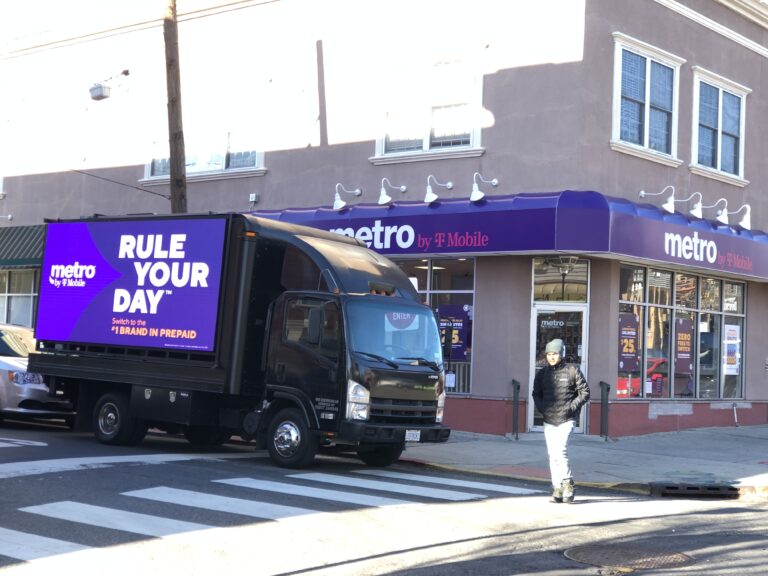 LED billboard truck for Metro by T-Mobile parked outside a store, featuring the slogan "RULE YOUR DAY" in bold letters, with a pedestrian in winter attire nearby.