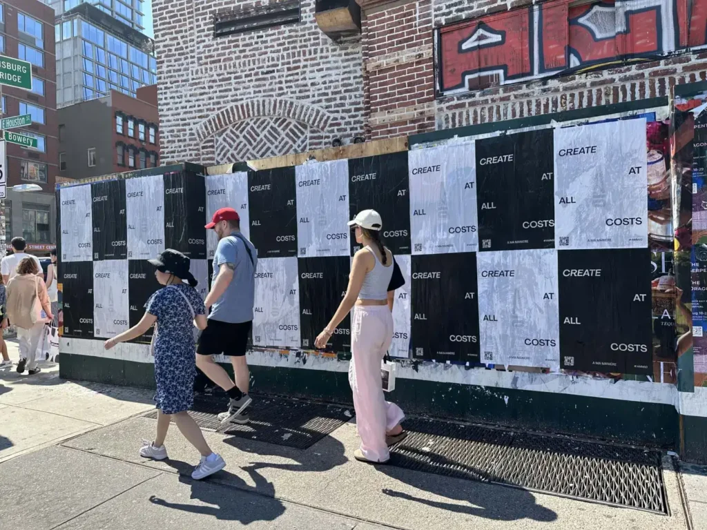 People walking past a wall covered in large black and white posters with the text "CREATE AT ALL COSTS," showcasing urban advertising and street marketing strategies.