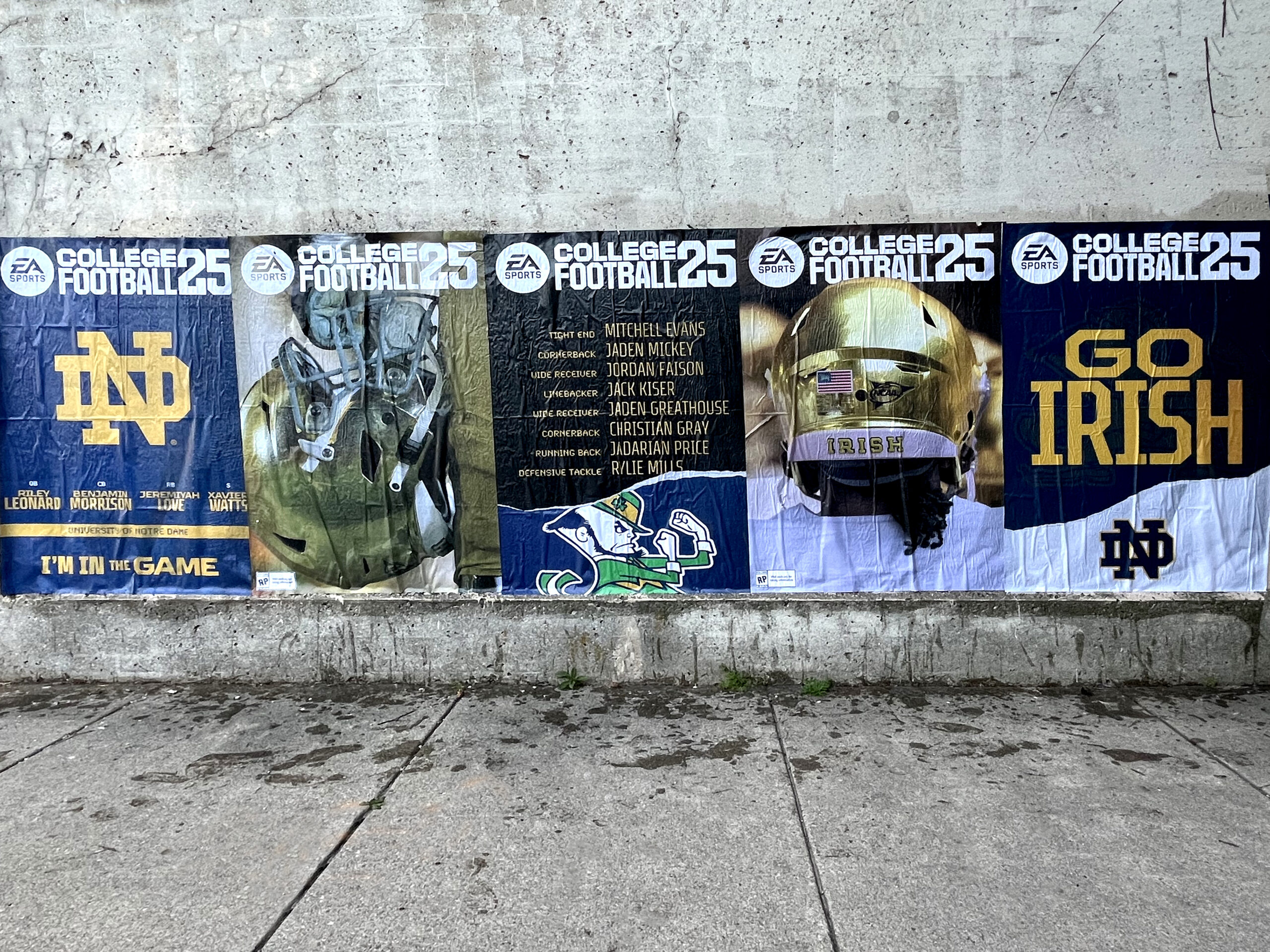 Wild wheat paste posting advertising featuring College Football 25 promotional banners, highlighting Notre Dame branding and player names, in an urban Indiana setting.
