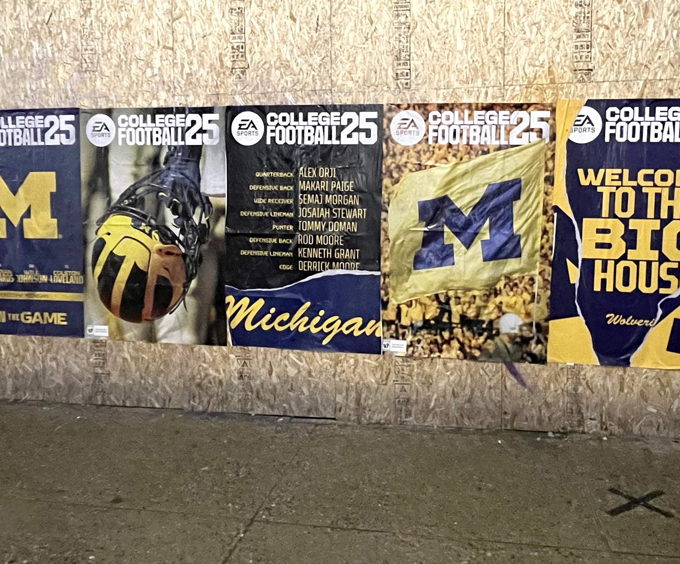 Posters featuring Michigan football themes, including a helmet, team logo, and reference to College Football 25, displayed on a wooden wall, highlighting urban advertising through wheat paste posting in Michigan.