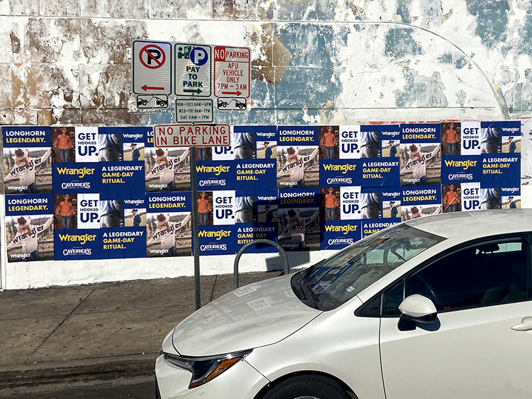 Wild wheat paste posting advertising in Minnesota featuring Wrangler promotional materials on a wall, with parking signs and a white car in the foreground.