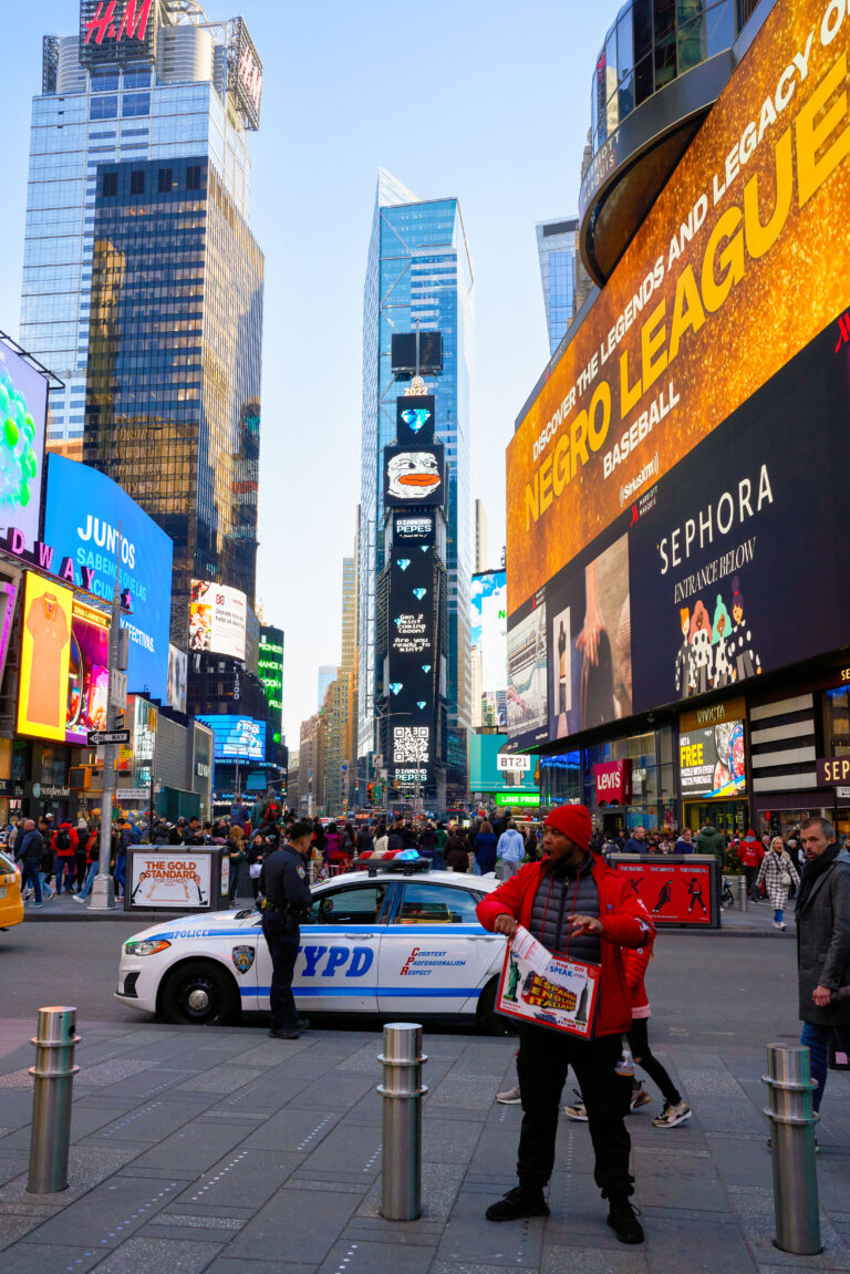 Crowded Times Square with a street vendor holding a sign, large digital billboards displaying advertisements, and an NYPD police car in the foreground, illustrating the vibrant atmosphere and high foot traffic relevant to the Diamond Peeps campaign.