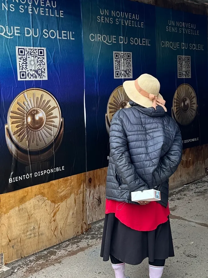 Woman in a hat standing in front of promotional posters for Cirque du Soleil, featuring a logo and QR codes, with text "Bient&ocirc;t Disponible" and "Un Nouveau Sens S'&Eacute;veille."
