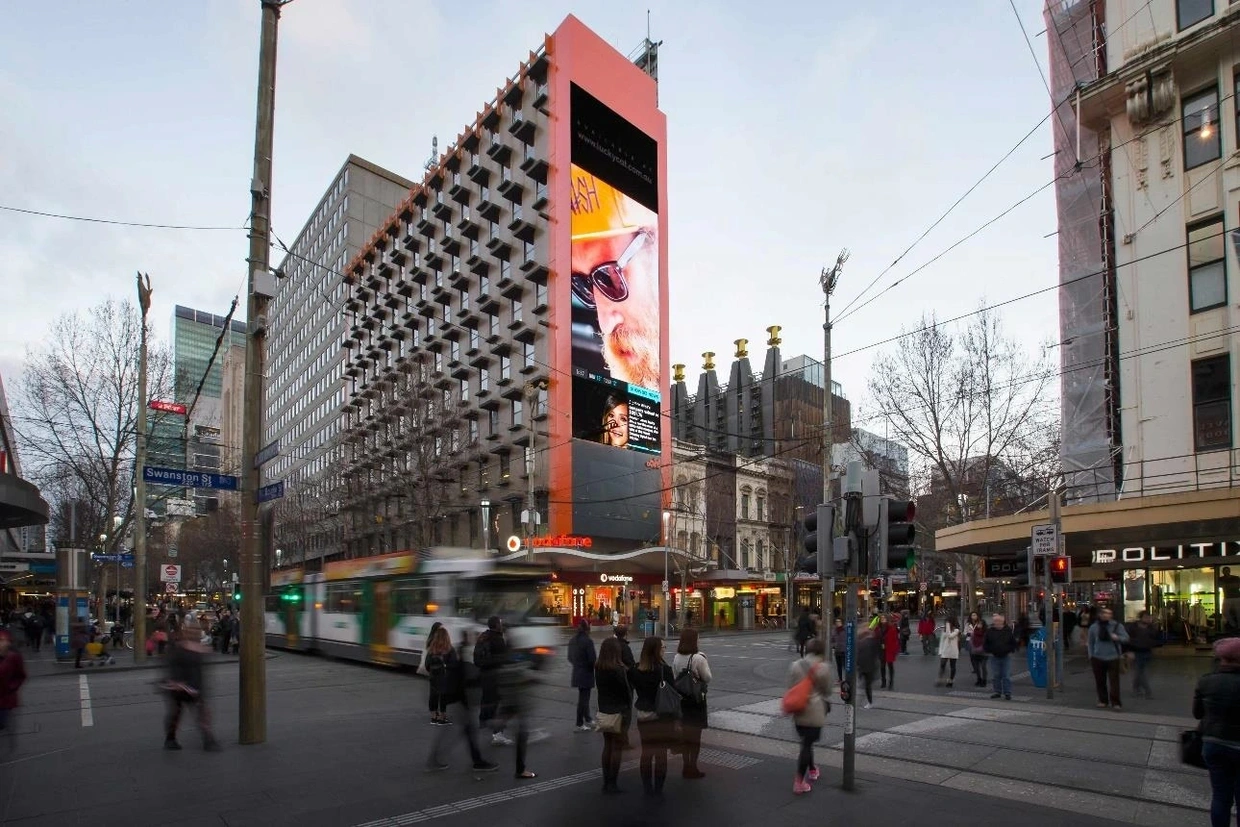 Urban scene featuring a large digital billboard on a building in a busy city intersection, showcasing advertising content, with pedestrians and a tram in motion, illustrating international billboard advertising potential.