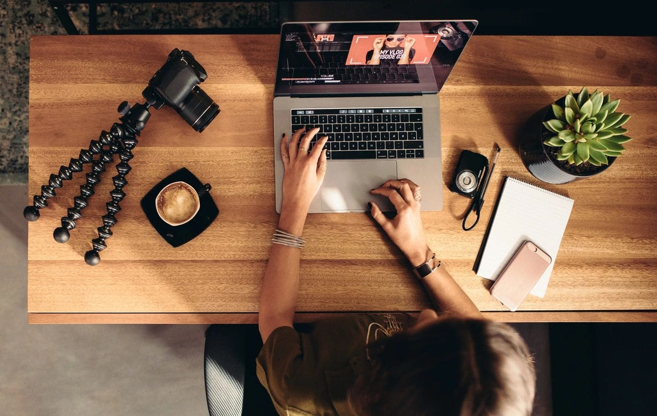 Person working on a laptop with camera, coffee, and notebook, illustrating content production and editing process for brand storytelling.