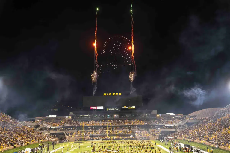 Drone light show with colorful aerial displays above a packed stadium at night, featuring fireworks and the word "MIZZOU" illuminated, enhancing the event atmosphere.