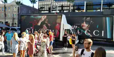 Static mobile vinyl billboard truck showcasing Gucci advertisement with a crowd of people in a busy urban setting.