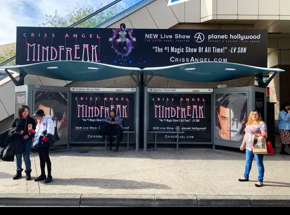 Bus stop advertising featuring Criss Angel's "Mindfreak" show, with commuters waiting at the shelter and promotional banners displaying show details and website information.