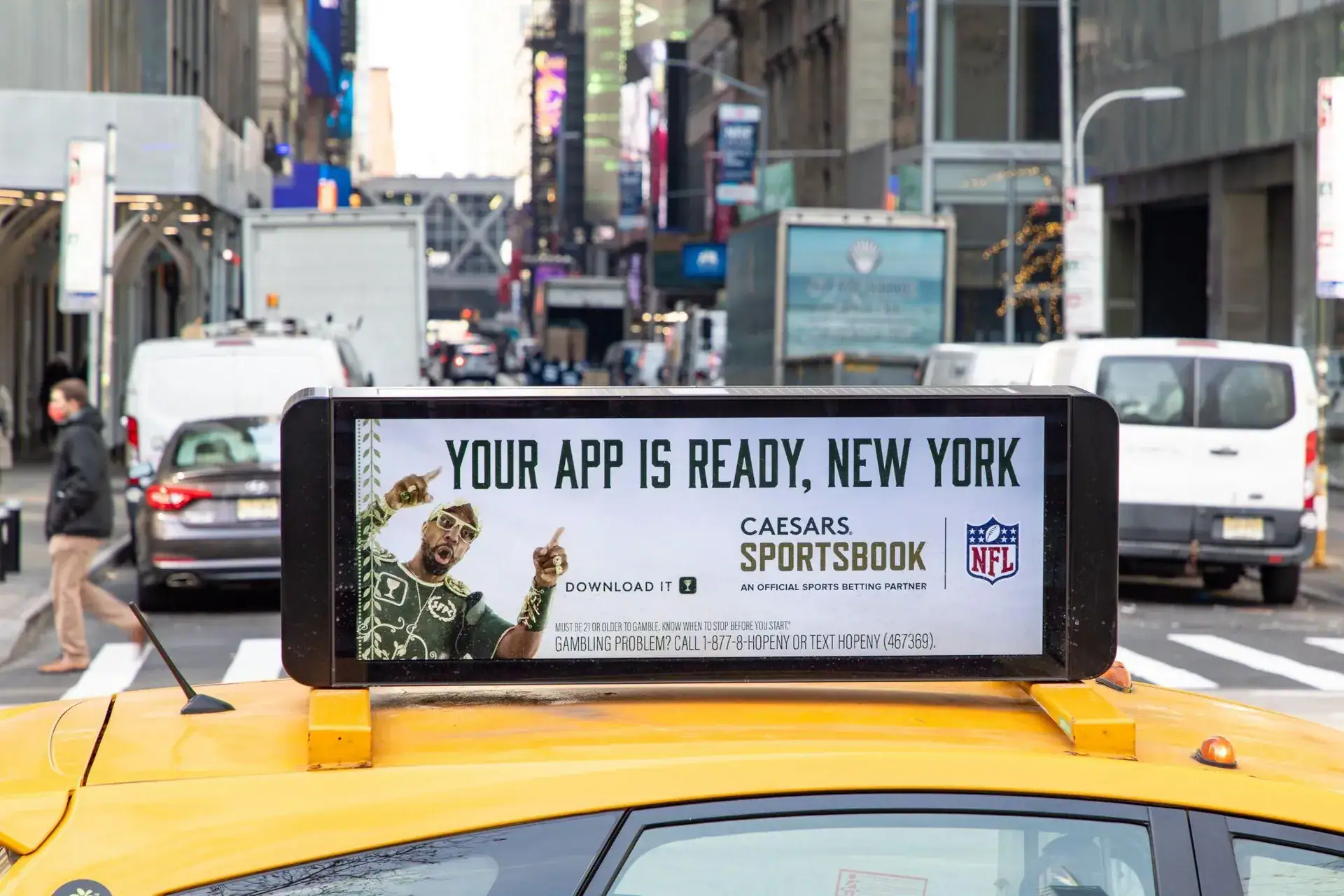 Taxi top advertisement featuring "Your App Is Ready, New York" for Caesars Sportsbook, with NFL branding, in a busy urban street setting.