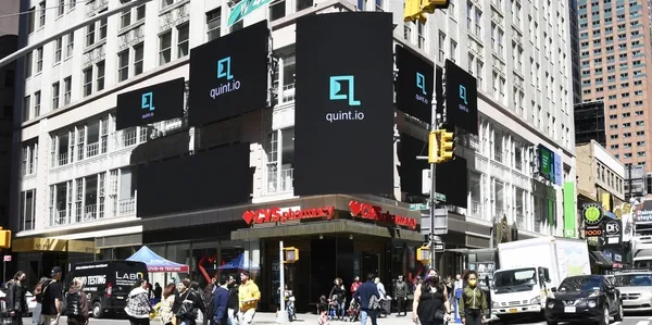 Billboard advertising featuring the logo of quint.io on a busy street corner, with pedestrians and vehicles in a vibrant urban setting, highlighting dynamic media visibility.