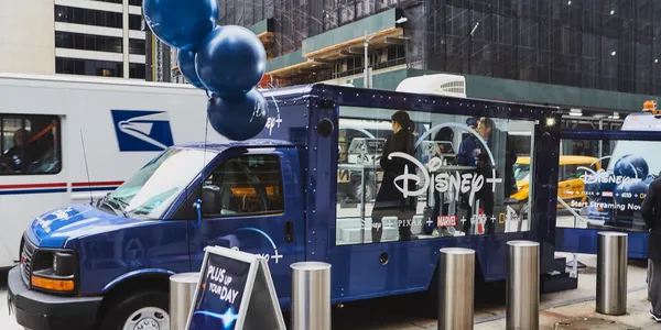 Disney+ promotional vehicle with blue balloons, showcasing streaming service branding, in an urban setting with pedestrians and vehicles nearby.