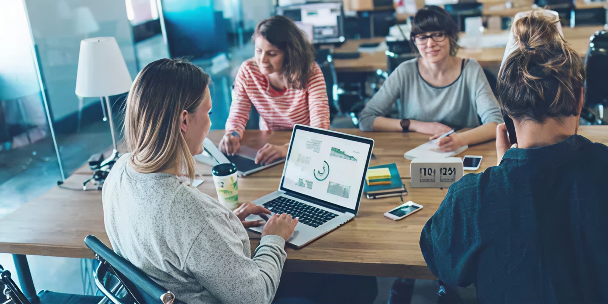 Group of four women collaborating at a workspace, one using a laptop displaying graphs, emphasizing teamwork and creativity in a professional setting.