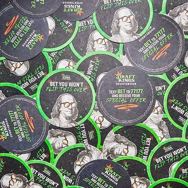 Coasters featuring DraftKings Sportsbook branding, promoting a special offer with text "Bet you won't flip this over" and instructions to text for more information, surrounded by a vibrant green and black design.