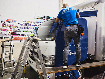 Person applying a blue wrap to a truck, showcasing a Disney logo, with ladders and design samples in the background, illustrating creative marketing execution for promotional visibility.