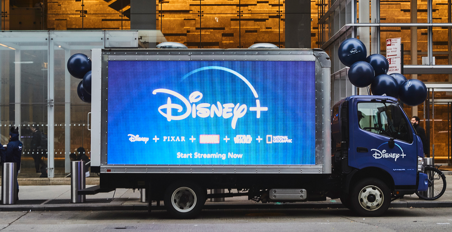 Disney+ promotional truck with vibrant blue display, showcasing Disney, Pixar, Marvel, and Star Wars logos, encouraging viewers to "Start Streaming Now," set against an urban backdrop with balloons.