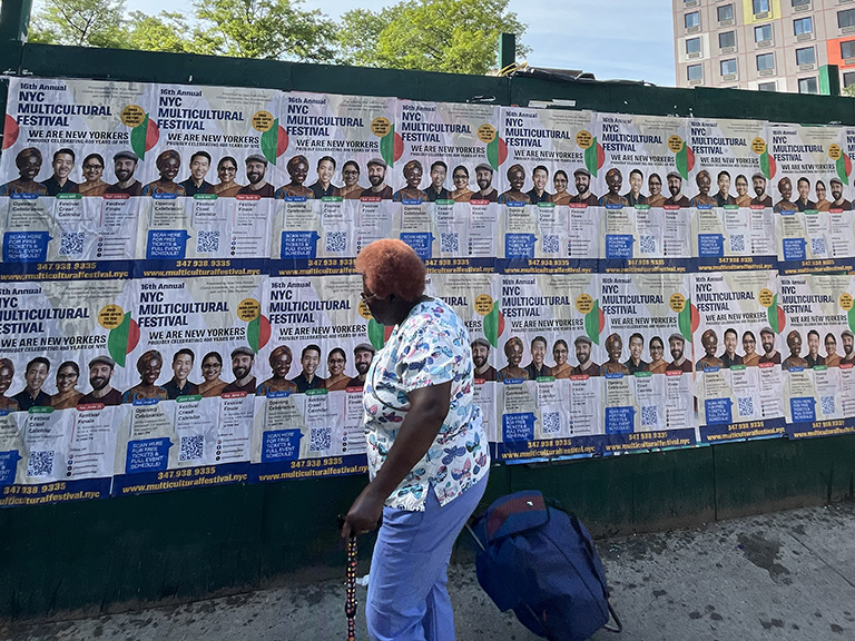 Woman walking past a wall covered with colorful posters for the NYC Multicultural Festival, featuring diverse individuals and event details, highlighting community-driven cultural engagement.