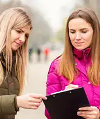 Two women engaged in a street survey, reviewing responses on a clipboard, with an urban backdrop, highlighting community engagement and feedback collection.