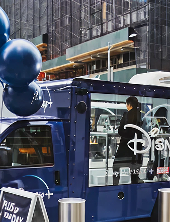 Disney+ promotional truck with balloons, featuring a woman inside, set against an urban backdrop, highlighting mobile fan experiences and brand visibility in high-traffic areas.