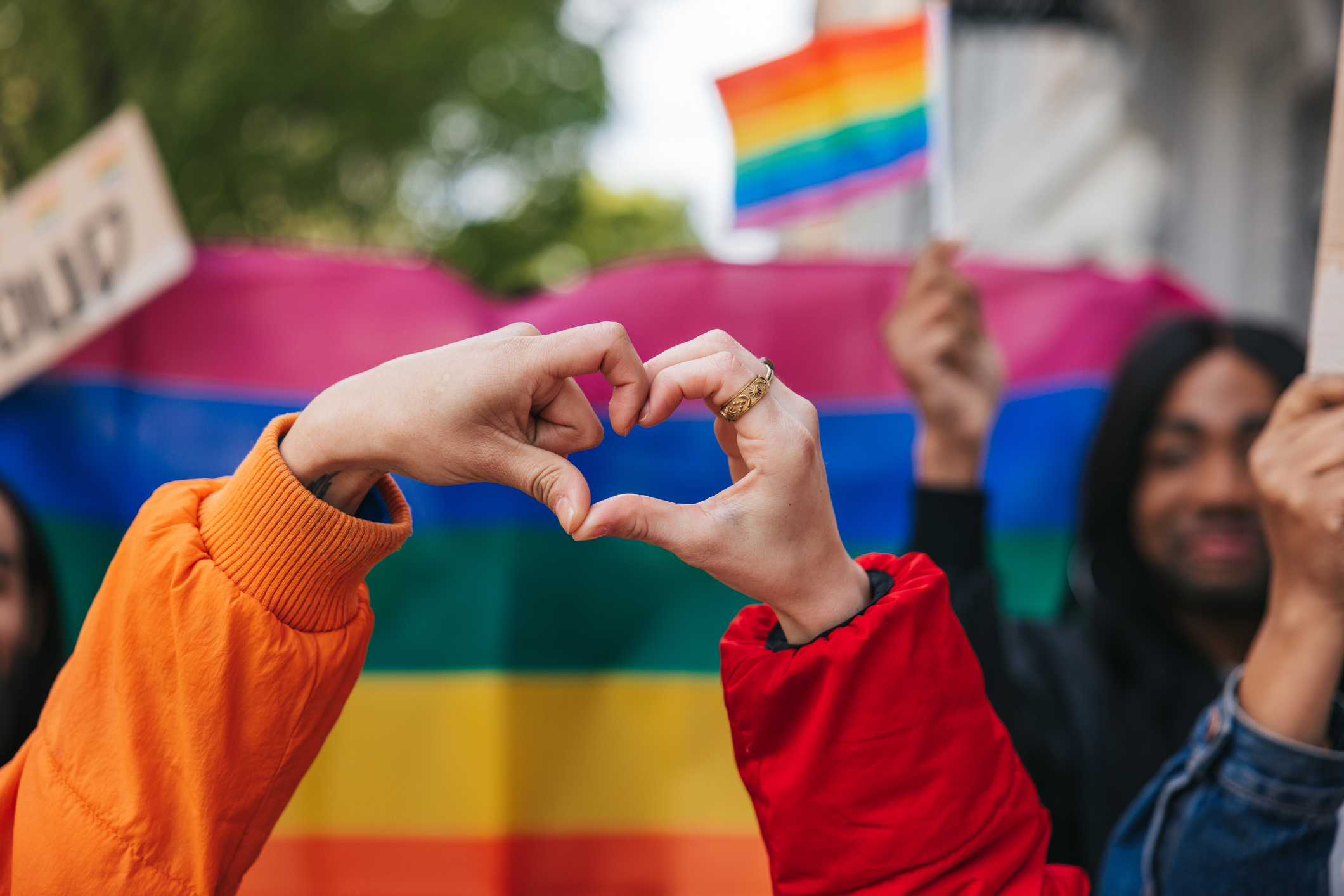 Hands forming a heart shape against a backdrop of a rainbow flag, symbolizing love and unity at a Pride event, with participants holding signs and flags celebrating LGBTQ+ identity and community engagement.
