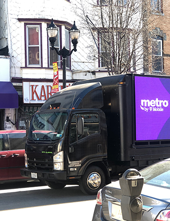 LED billboard truck displaying Metro by T-Mobile advertisement, parked on a city street near local businesses, emphasizing mobile marketing visibility in urban environments.