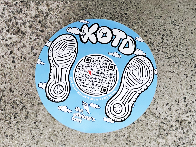 Sidewalk decal for The Athlete&rsquo;s Foot featuring "KOTD" text, QR code for discounts, and sneaker footprints on a blue background, promoting a street marketing campaign.