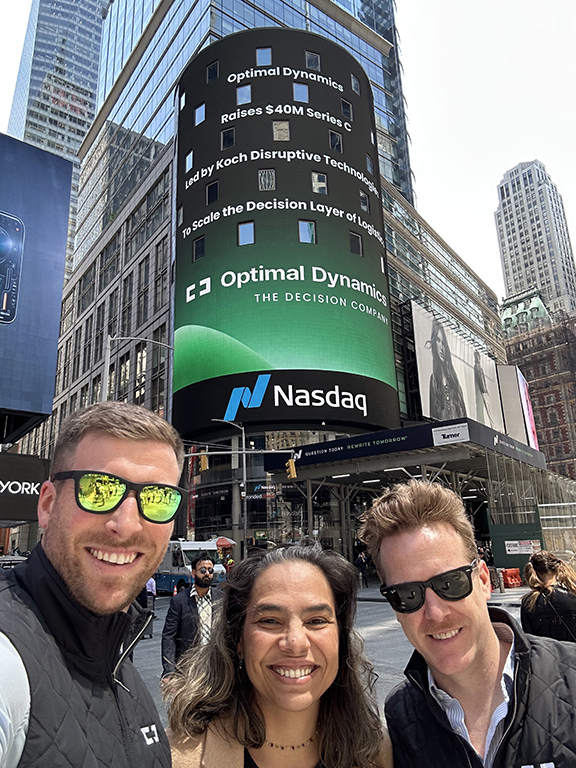 Smiling group of three people in front of NASDAQ digital billboard displaying Optimal Dynamics advertising campaign in Times Square.