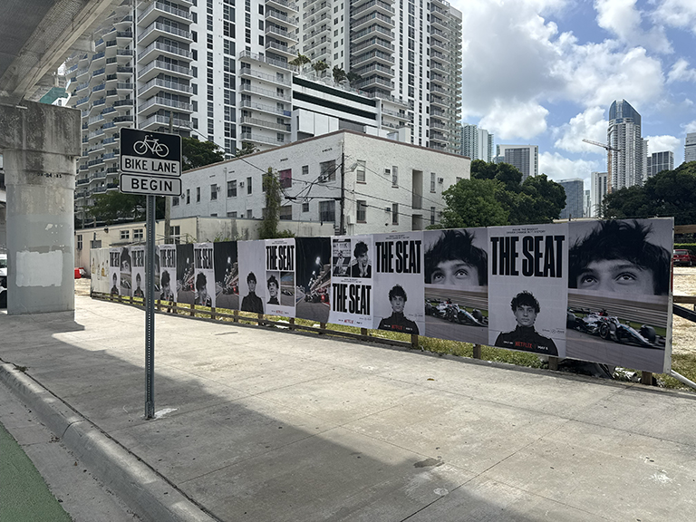Billboard advertising for Netflix's "The Seat" featuring multiple large posters along a city street, with urban buildings in the background and a bike lane sign.