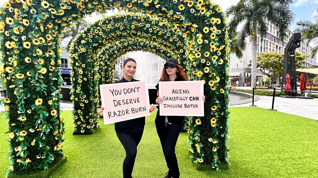 Two women holding signs promoting skincare and beauty messages, standing under a floral arch in a vibrant urban setting, with greenery and palm trees in the background.