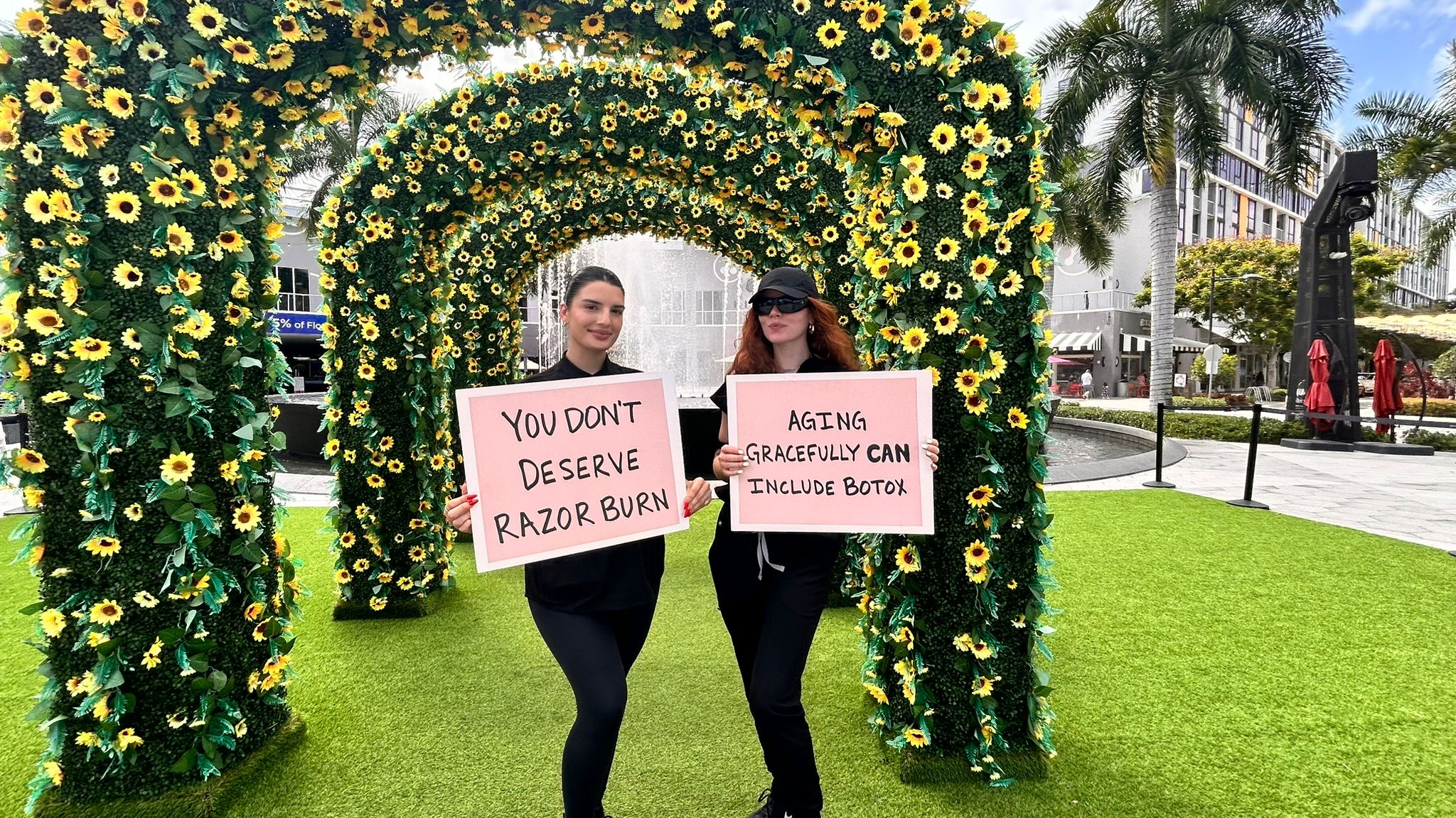 Two brand ambassadors holding signs with messages about skincare and aging, standing under a floral arch at a promotional event in a vibrant outdoor setting.