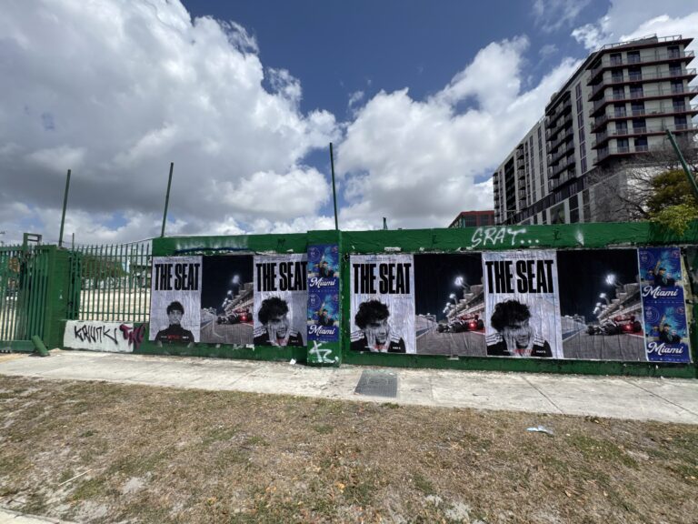 Wheatpasted posters promoting Netflix's "The Seat" campaign, featuring a cinematic design with bold text, installed on a green wall in Miami, surrounded by urban elements and graffiti, capturing the vibrant atmosphere during the F1 Grand Prix weekend.