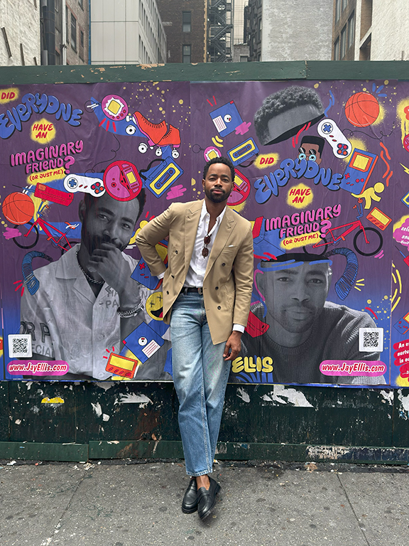 Jay Ellis posing in front of a colorful wheatpaste poster promoting his book, featuring playful graphics and text about imaginary friends, in an urban setting of Manhattan.