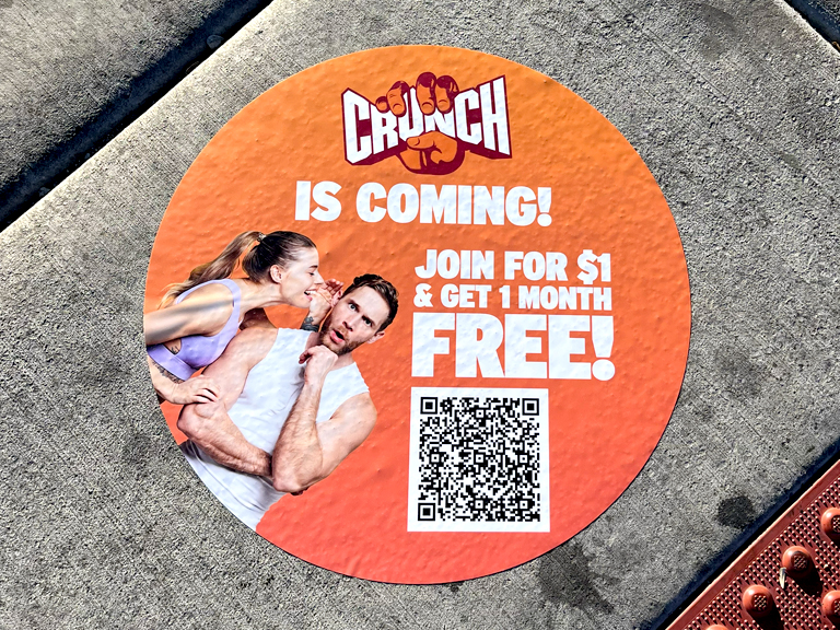 Crunch Fitness sidewalk decal advertising campaign, featuring vibrant orange background, promotional text "CRUNCH IS COMING! JOIN FOR $1 & GET 1 MONTH FREE!" with images of fitness enthusiasts and a QR code.