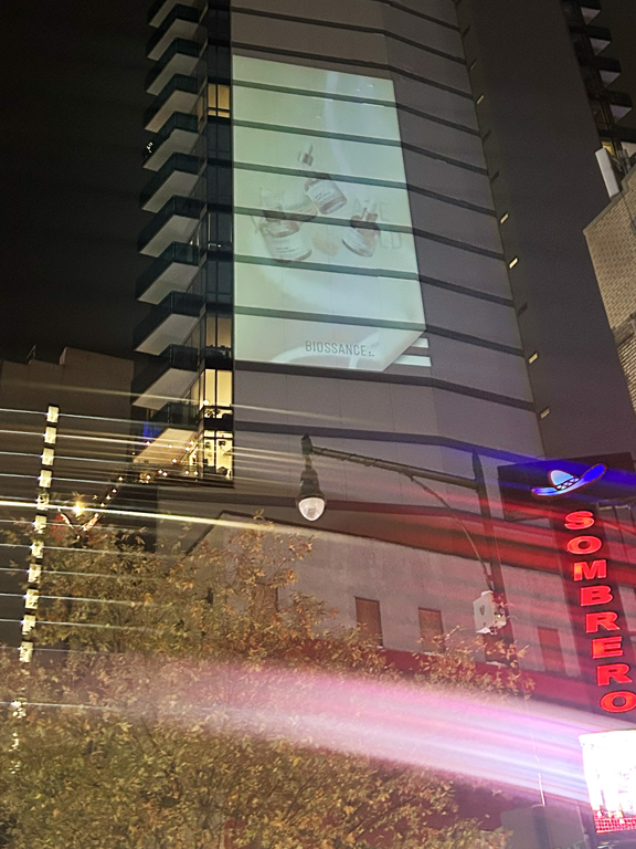Billboard advertisement for Biossance displayed on a high-rise building at night, featuring skincare products, with surrounding urban elements including streetlights and a nearby restaurant sign.