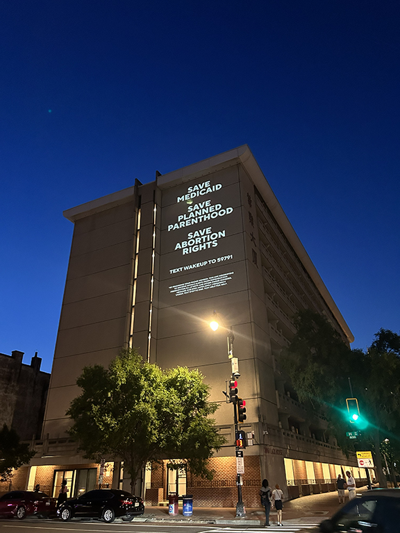 Projection of advocacy messages on a building, promoting healthcare and abortion rights, featuring text "Save Medicaid," "Save Planned Parenthood," and "Save Abortion Rights," illuminated at night.