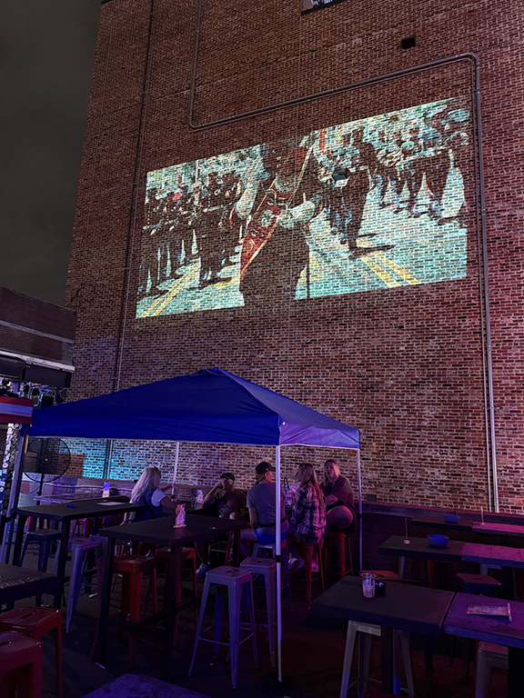 Projection of a marching band on a brick wall, with a group of people sitting under a tent in a dimly lit outdoor setting, showcasing guerrilla projection advertising.