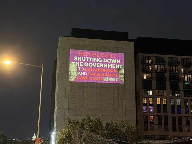 Projection of a political message on a building, stating "REPUBLICANS: SHUTTING DOWN THE GOVERNMENT TO BAN ABORTION AND RAISE HEALTH CARE COSTS," part of an advocacy projection campaign for reproductive freedom.