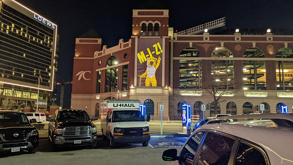 Mizzou-themed guerrilla projection campaign featuring a large, colorful banner of a mascot with the text "M-I-Z!" on a building at night, surrounded by parked vehicles in Arlington, Texas.
