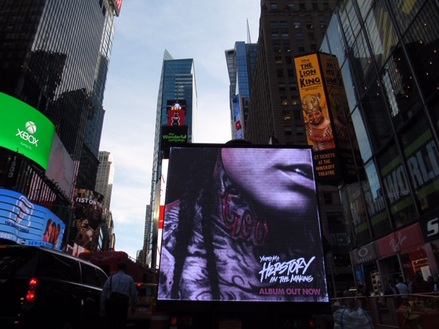 14ft LED billboard truck displaying Young M.A's "Herstory in the Making" album advertisement in Times Square, New York, surrounded by urban skyscrapers and other digital advertisements.