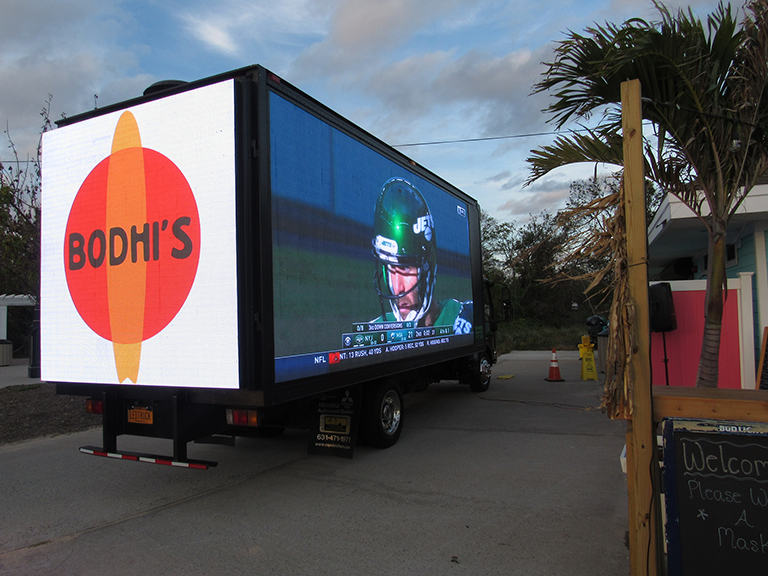14ft LED billboard truck displaying "BODHI'S" logo and live sports broadcast, positioned in a vibrant outdoor setting, showcasing mobile advertising for Bodhi's in Montauk, New York.