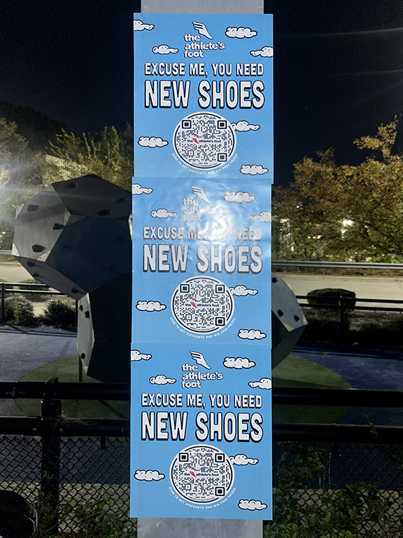 Promotional poster for The Athlete&rsquo;s Foot featuring the slogan "Excuse Me, You Need New Shoes" and a QR code, displayed at night with a park setting in the background.