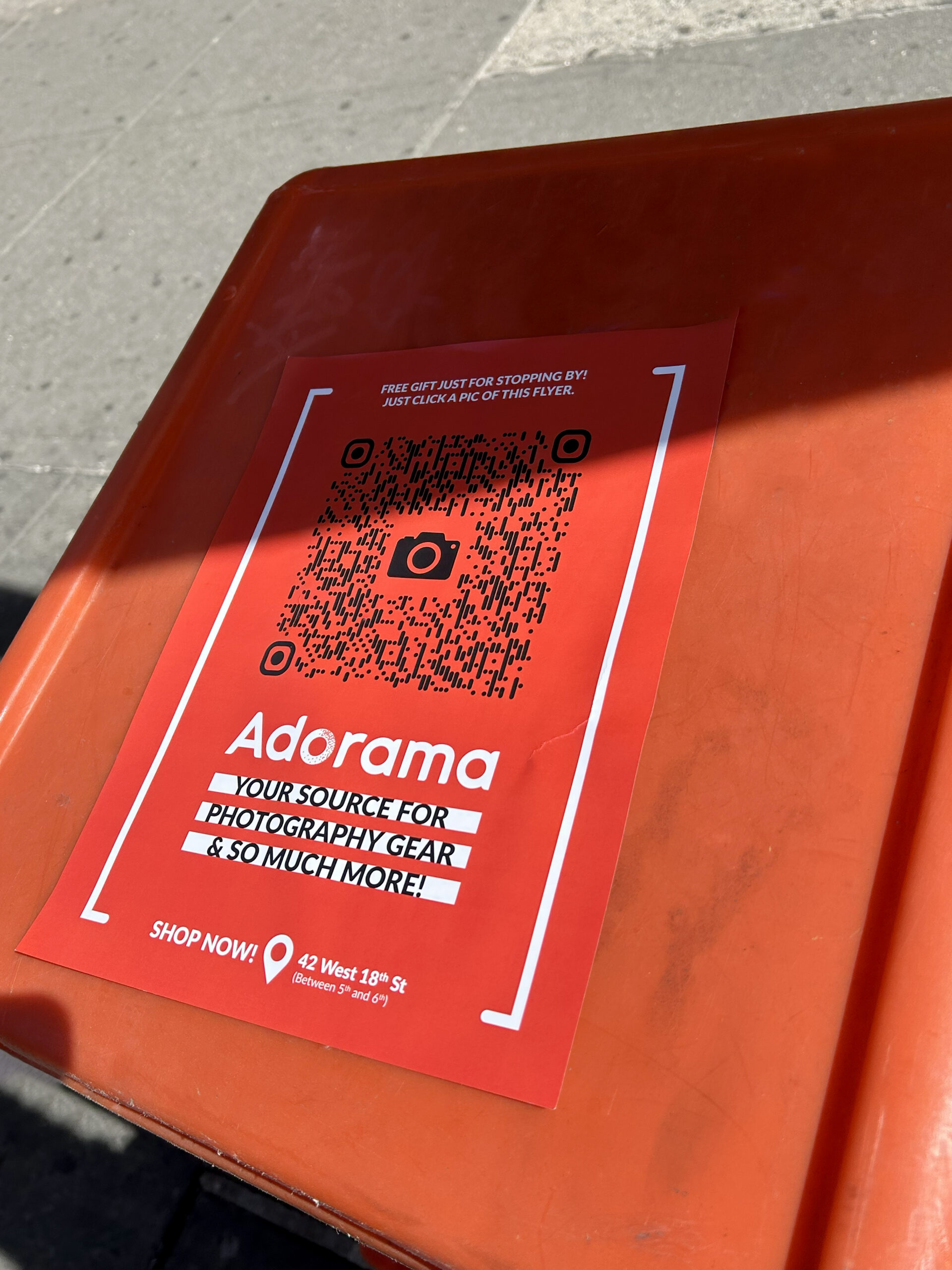 Red poster advertising Adorama with QR code, promoting free gift for in-store visit, highlighting photography gear and services, placed on an orange surface.