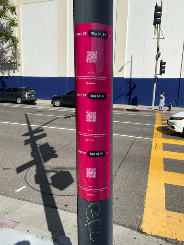 Pole displaying multiple pink advertisement posters for Vivira's "Tired Of Ads? Skip Ad" campaign, featuring a QR code and engaging call-to-action, showcasing guerrilla marketing techniques in urban Los Angeles.
