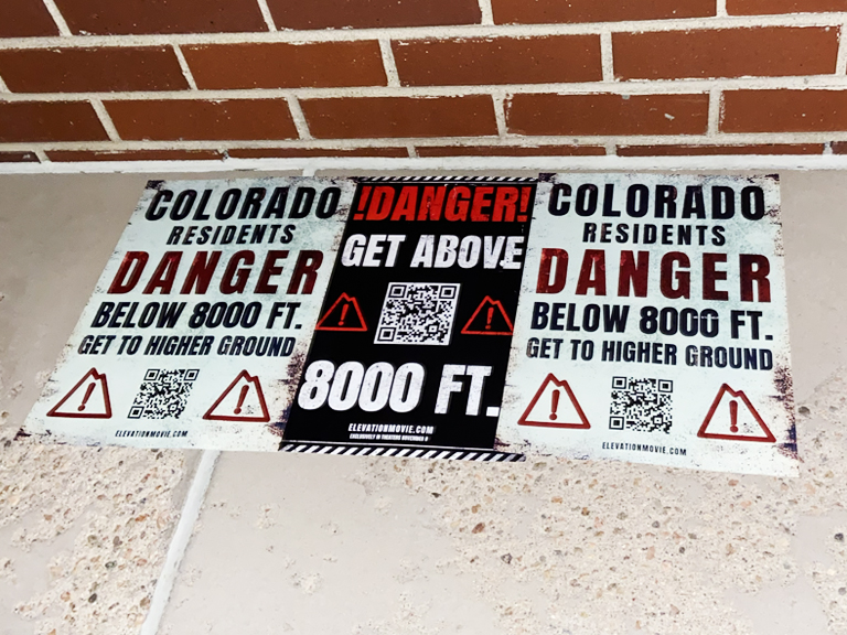Signs displaying warnings for Colorado residents about danger below 8000 feet, featuring QR codes and instructions to get to higher ground, emphasizing safety for the Elevation movie promotion.