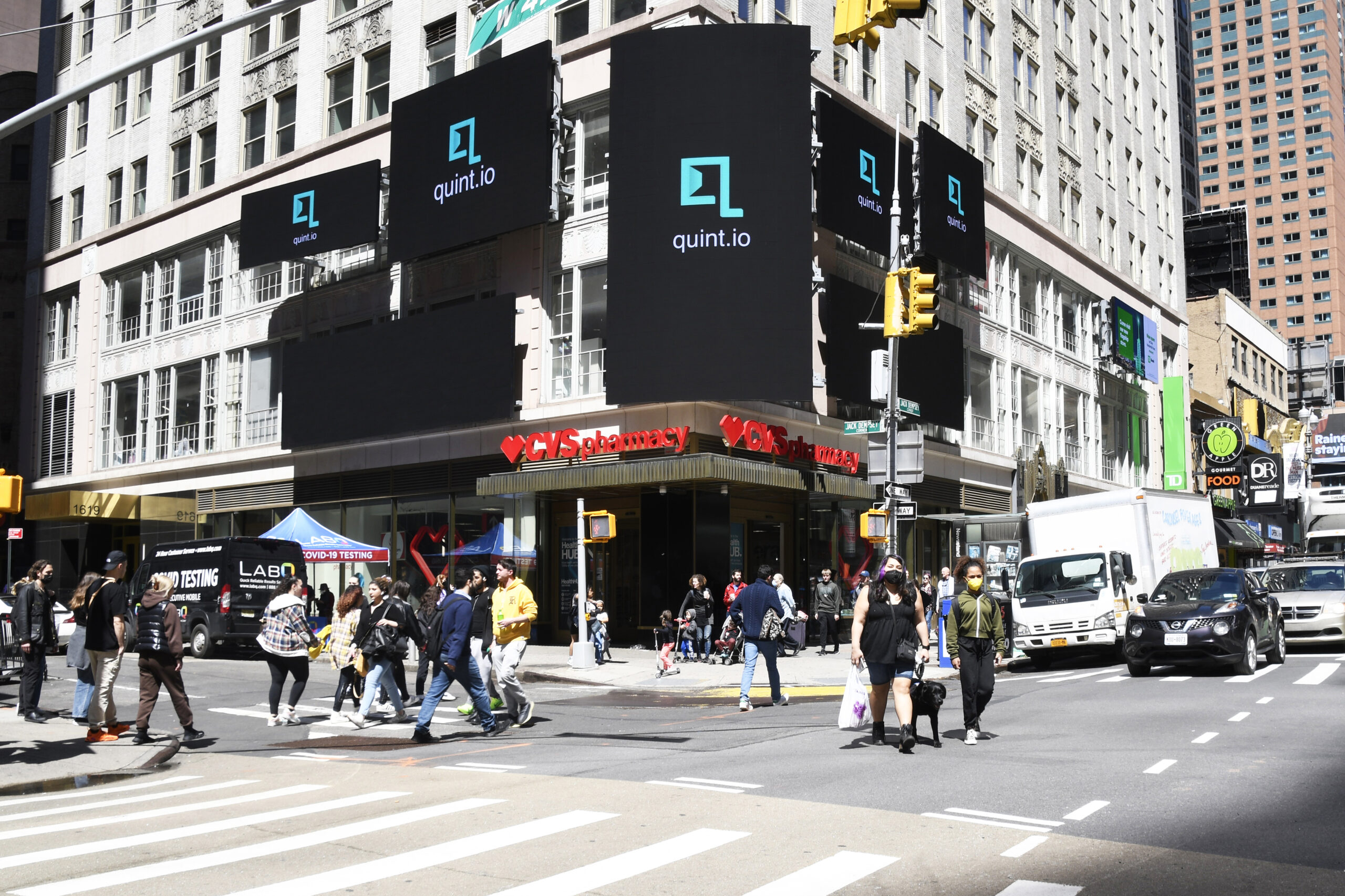 Quint.io digital billboards prominently displayed on the Brill Building in Times Square, New York City, with pedestrians crossing the street below.
