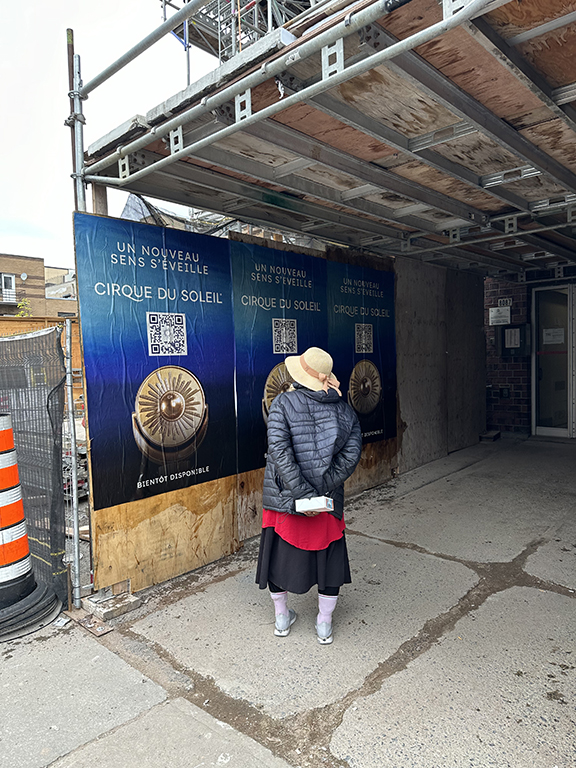 Woman in a black jacket and hat standing in front of Cirque du Soleil wheatpaste posters advertising "Un Nouveau Sens S'&eacute;veille" with QR codes, in an urban construction setting.